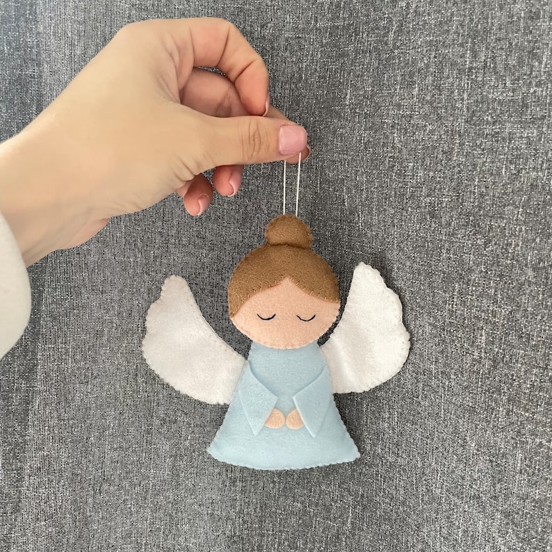 Puede incluir: Un adorno de &aacute;ngel de fieltro con alas blancas, cabello casta&ntilde;o y un vestido azul claro. El &aacute;ngel tiene los ojos cerrados y las manos juntas en oraci&oacute;n.