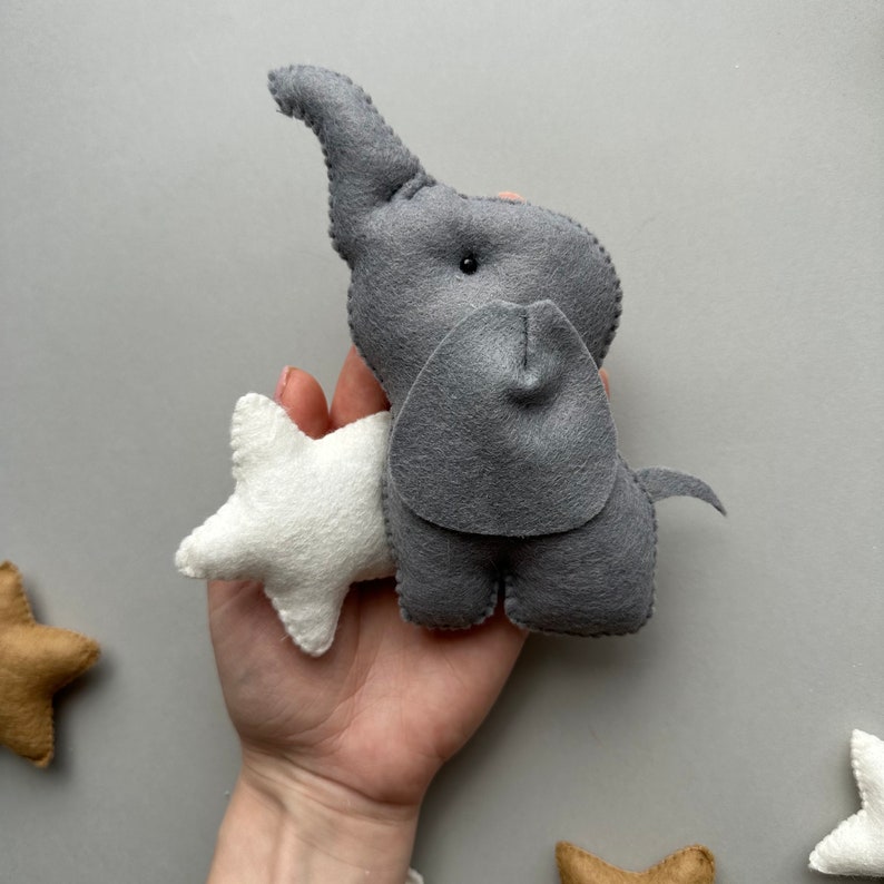 May include: A grey felt elephant toy with a white felt star attached to its trunk. The elephant has a small black eye and a grey felt trunk.