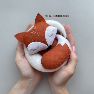 May include: A felt fox plush toy with closed eyes, curled up in a sleeping position. The fox is orange and white with embroidered details. The text "PDF PATTERN FOX-MOON" is visible in the image.