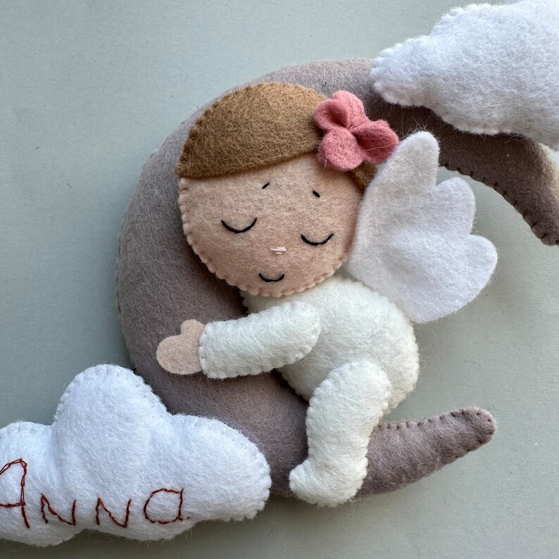 Pu&ograve; includere: Un angelo di feltro con capelli castani e un fiore rosa che dorme su una luna crescente grigia. L'angelo indossa una tutina bianca e ha ali bianche. La luna &egrave; circondata da due nuvole bianche, una delle quali ha il nome "Anna" ricamato.