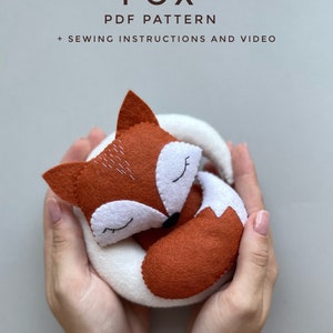 以下が含まれることがあります： かわいい丸まったキツネのぬいぐるみのPDF縫製パターンです。パターンには縫製手順とビデオチュートリアルが含まれています。キツネはフェルト製で、白いお腹と茶色の体を持っています。キツネは手のひらに乗っています。