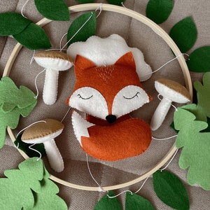 May include: A wooden hoop with a felt fox, mushrooms, and green leaves hanging from it. The fox is orange and white with closed eyes. The mushrooms are brown and white. The leaves are a deep green.