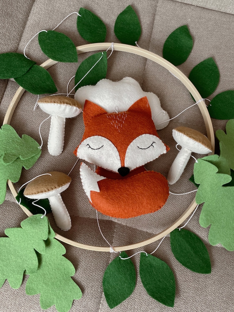 May include: A wooden hoop with a felt fox, mushrooms, and green leaves hanging from it. The fox is orange and white with closed eyes. The mushrooms are brown and white. The leaves are a deep green.
