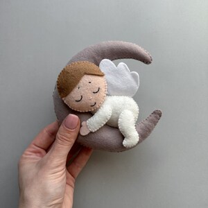 Pu&ograve; includere: Un angelo di feltro con capelli castani e occhi chiusi dorme su una luna crescente grigia. L'angelo indossa un abito di feltro bianco con un'ala di feltro bianco.