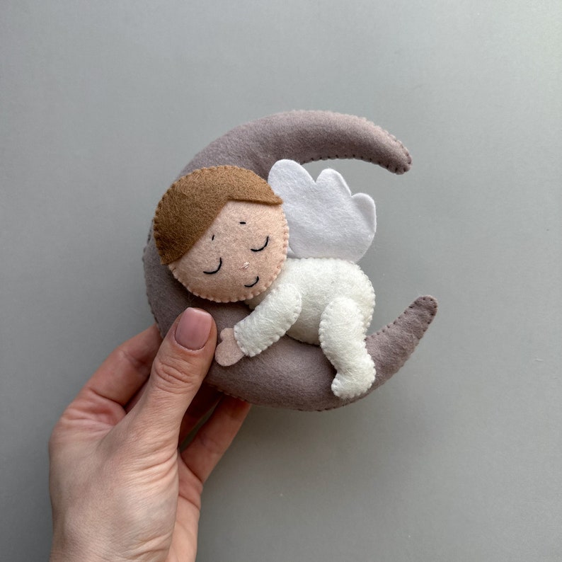 Pu&ograve; includere: Un angelo di feltro con capelli castani e occhi chiusi dorme su una luna crescente grigia. L'angelo indossa un abito di feltro bianco con un'ala di feltro bianco.