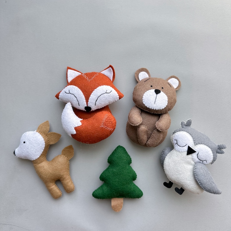 May include: Five felt animal toys: a brown deer, a red fox, a brown bear, a gray owl, and a green tree. The toys are all handmade and have a simple, whimsical design.
