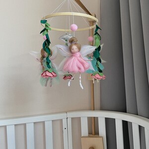 May include: A wooden baby mobile with a circular frame and five hanging figures. The figures are made of felt and include fairies with pink and green dresses, white wings, and pink flowers. There are also green leaves and pink pom-poms hanging from the frame.