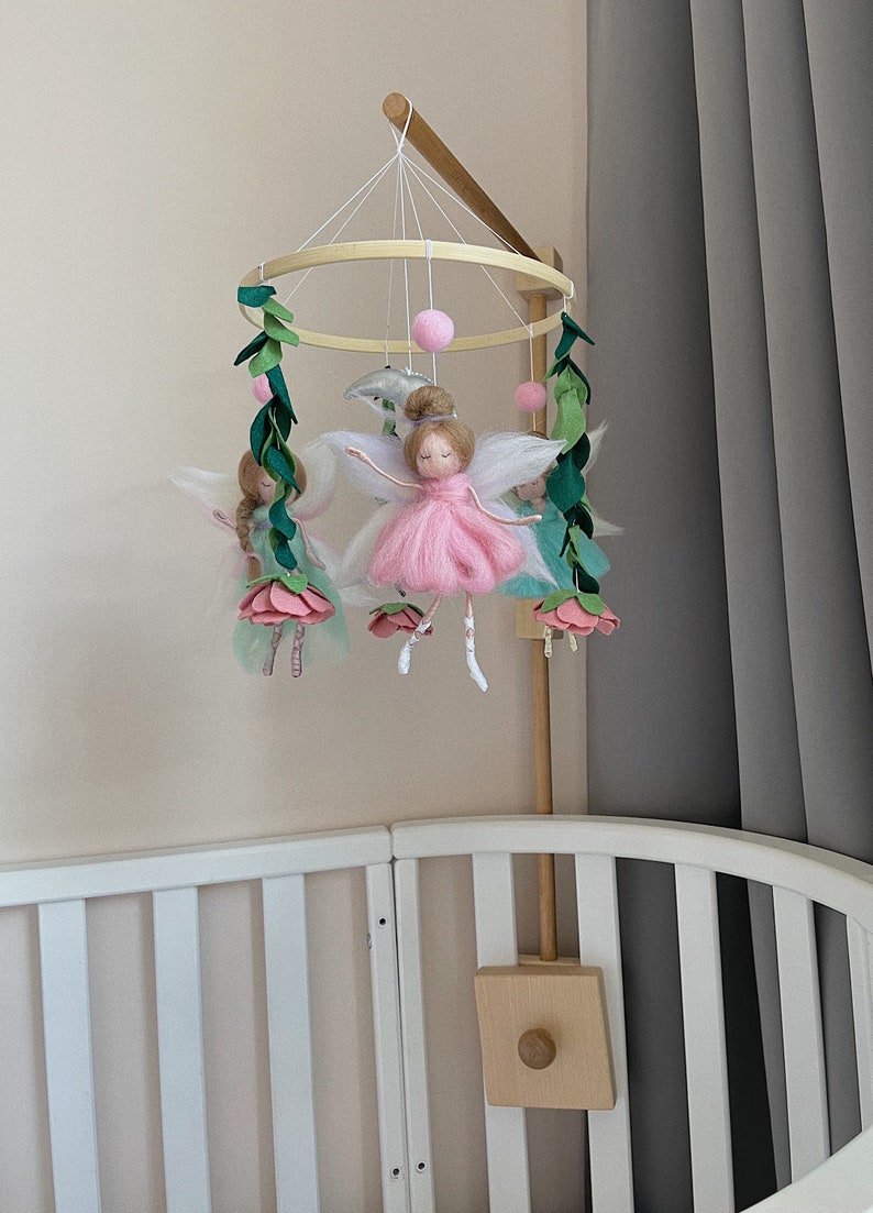 May include: A wooden baby mobile with a circular frame and five hanging figures. The figures are made of felt and include fairies with pink and green dresses, white wings, and pink flowers. There are also green leaves and pink pom-poms hanging from the frame.