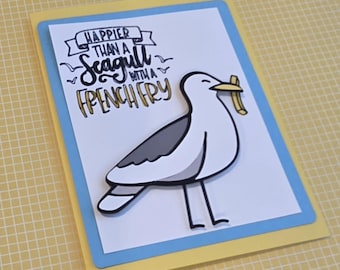 Puede incluir: Una tarjeta de felicitación rectangular con una gaviota de dibujos animados que sostiene una patata frita amarilla en el pico. El texto dice HAPPIER THAN A SEAGULL WITH A FRENCH FRY. La tarjeta tiene un centro blanco, un borde azul claro y una base amarilla.