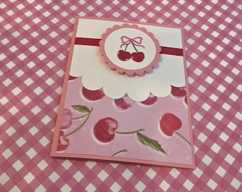 Puede incluir: Una tarjeta de felicitación rectangular con fondo rosa claro y un patrón repetitivo de cerezas. La parte superior tiene un borde festoneado blanco, una franja horizontal rosa oscuro y un medallón circular con un gráfico de cerezas.