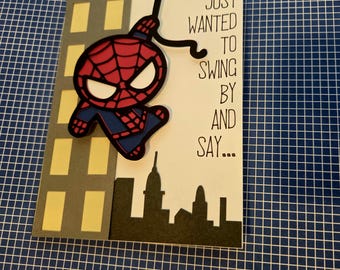 Puede incluir: Una tarjeta de felicitación con un personaje de superhéroe rojo y azul troquelado balanceándose en una telaraña negra. La tarjeta incluye un edificio gris con ventanas amarillas, una silueta negra de la ciudad y el texto: JUST WANTED TO SWING BY AND SAY...