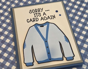 Puede incluir: Una tarjeta de felicitación rectangular con la ilustración de una rebeca azul claro con ribetes y botones azul oscuro. El texto SORRY... IT'S A CARD AGAIN está impreso en tinta negra sobre la rebeca. La tarjeta tiene un centro blanco, un borde gris y un borde exterior azul oscuro.