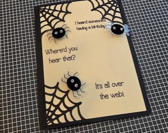 Puede incluir: Una tarjeta de felicitación rectangular con un centro color crema y bordes negros que presentan telarañas troqueladas en las esquinas. Tres arañas negras con ojos móviles están adheridas. El texto dice: I heard someone's having a birthday, Where'd you hear that?, It's all over the web!