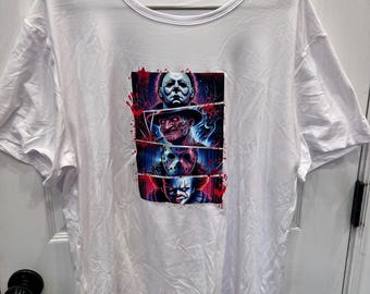 Puede incluir: Una camiseta blanca de manga corta con un estampado gráfico central dividido en cuatro paneles horizontales. La ilustración muestra cuatro personajes de películas de terror distintos con salpicaduras de sangre roja sobre un fondo azul y morado vibrante.
