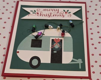 Puede incluir: Una tarjeta de felicitación hecha a mano con una caravana vintage de color turquesa y blanco sobre un fondo verde oscuro. La tarjeta muestra una pancarta con el texto merry christmas y una cadena de mini bombillas navideñas.