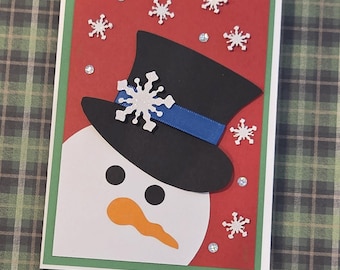 Puede incluir: Una tarjeta de felicitación hecha a mano con la cara de un muñeco de nieve, cabeza blanca, ojos circulares negros y nariz de zanahoria naranja. Lleva un sombrero de copa negro con una cinta azul y un copo de nieve blanco.