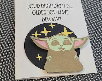 Puede incluir: Una tarjeta de felicitación blanca con un fondo circular negro con estrellas amarillas y un recorte de papel en capas de una criatura verde con orejas grandes. El texto en la tarjeta dice: YOUR BIRTHDAY IT IS.... OLDER YOU HAVE BECOME!