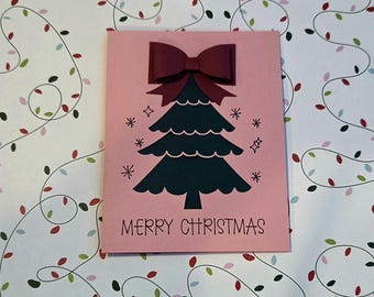Puede incluir: Una tarjeta de felicitación de color rosa claro con una ilustración de un árbol de Navidad verde oscuro coronado con un lazo de papel rojo oscuro. La tarjeta incluye estrellas negras y el texto MERRY CHRISTMAS en una fuente sencilla.