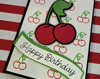 Puede incluir: Una tarjeta de felicitación rectangular con fondo blanco que presenta un patrón repetitivo de pequeñas cerezas rojas con tallos verdes. Un par de cerezas troqueladas se sitúa en la parte superior y una cinta con borde verde muestra el texto Happy Birthday.