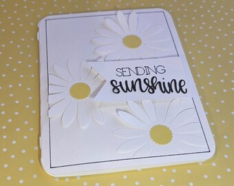 Puede incluir: Una tarjeta de felicitación rectangular blanca con tres margaritas blancas troqueladas con centros amarillos. Una banda blanca se superpone en el centro, impresa con el texto negro "SENDING sunshine" en una combinación de fuentes serif y manuscrita.