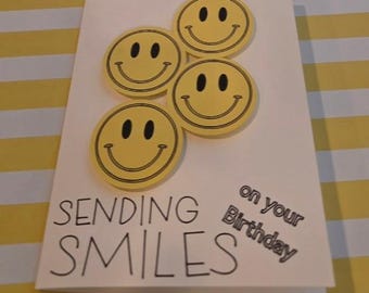 Puede incluir: Una tarjeta de felicitación blanca con tres iconos circulares amarillos de caras sonrientes con contornos y rasgos faciales negros. El texto de la tarjeta dice "SENDING SMILES on your Birthday" en letras negras con una fuente clara y legible.