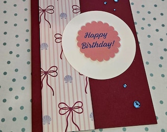 Puede incluir: Una tarjeta de felicitación granate con un panel vertical de rayas rosa claro, lazos de color rojo oscuro y pequeñas conchas de color violeta claro. Un círculo blanco contiene un centro rosa festoneado con el texto azul Happy Birthday y pedrería azul.