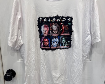 Puede incluir: Una camiseta blanca de manga corta con un estampado gráfico de seis personajes de películas de terror en una cuadrícula. El texto "F.R.I.E.N.D.S" aparece en la parte superior, con los nombres y lugares debajo de cada figura en tinta negra.