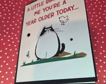 Puede incluir: Una tarjeta de felicitación blanca con borde negro que presenta una ilustración de dibujos animados de un gato redondo blanco y negro, un pájaro pequeño y una mariposa. El texto dice: A LITTLE BIRDIE TOLD ME YOU'RE A YEAR OLDER TODAY... con tres puntos negros.