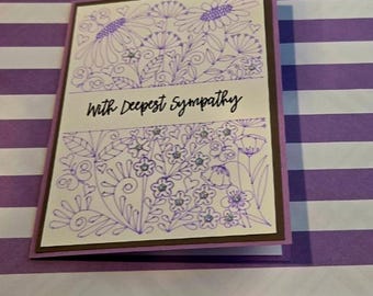 Puede incluir: Una tarjeta de condolencias rectangular con un panel frontal blanco que presenta dibujos de líneas moradas de varias flores, hojas y corazones pequeños. El texto "With Deepest Sympathy" está impreso en negro en el centro. Borde marrón oscuro.