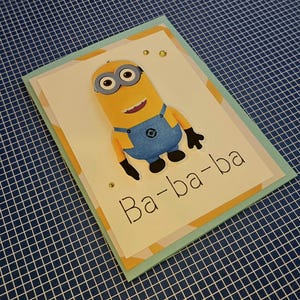 Puede incluir: Una tarjeta de felicitación rectangular que presenta un personaje Minion amarillo con peto azul. La tarjeta tiene un fondo blanco con el texto Ba-ba-ba impreso en negro. El diseño incluye pequeños adornos dorados y un borde azul claro.