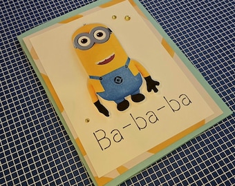 Puede incluir: Una tarjeta de felicitación rectangular que presenta un personaje Minion amarillo con peto azul. La tarjeta tiene un fondo blanco con el texto Ba-ba-ba impreso en negro. El diseño incluye pequeños adornos dorados y un borde azul claro.