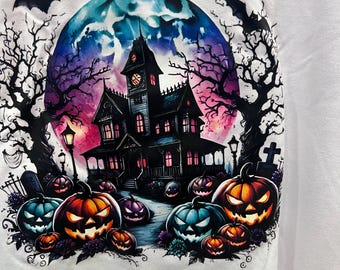 Puede incluir: Una camiseta blanca con un estampado gráfico de una casa encantada bajo una gran luna azul y morada. La escena incluye siluetas de árboles desnudos, murciélagos volando y varias calabazas brillantes con caras talladas en naranja y azul.