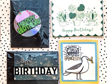 Puede incluir: Cuatro tarjetas de felicitación en fundas de plástico transparente. Una presenta un círculo holográfico con texto verde que dice Happy Birthday. Otra muestra plantas dibujadas con líneas verdes y el texto Happy Birthday. Una tercera tiene un fondo estrellado con el texto happy BIRTHDAY. La cuarta muestra una gaviota con una patata frita y el texto Happier than a seagull with a french fry.
