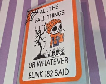 Puede incluir: Una tarjeta de felicitación con borde naranja que muestra un esqueleto con gorro y bufanda naranja, sosteniendo una bebida. El texto dice ALL THE FALL THINGS OR WHATEVER BLINK 182 SAID. Una telaraña negra está en la esquina superior izquierda.
