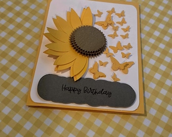 Puede incluir: Una tarjeta de felicitación rectangular con fondo blanco que presenta un girasol de papel amarillo en capas con un centro oscuro. Numerosas mariposas de papel amarillas pequeñas están dispuestas a la derecha de la flor. Una etiqueta festoneada gris oscuro con el texto Happy Birthday se encuentra en la parte inferior.
