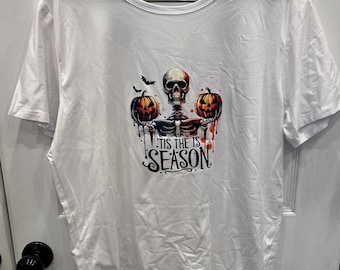 Puede incluir: Una camiseta blanca de manga corta con un estampado gráfico de un esqueleto que sostiene dos calabazas de Halloween naranjas. Sobre el esqueleto hay dos pequeños murciélagos negros. Debajo, el texto dice 'TIS THE IS SEASON' en letras negras.