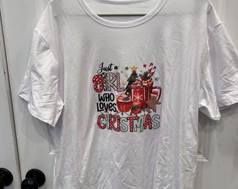 Puede incluir: Una camiseta blanca de manga corta con un estampado gráfico colorido que incluye el texto "Just a GIRL WHO LOVES CRISTMAS". El diseño muestra una taza roja, un cupcake, un hombre de jengibre, un árbol de Navidad y bastones de caramelo.