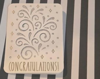 Puede incluir: Una tarjeta de felicitación rectangular blanca con un diseño de fuegos artificiales o flores con purpurina plateada, junto a pequeñas estrellas y puntos. La palabra CONGRATULATIONS! está impresa en la parte inferior en letra sencilla.