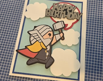 Puede incluir: Una tarjeta de felicitación rectangular con un personaje de dibujos animados que lleva un casco plateado y una capa roja y amarilla, sosteniendo un martillo gris. La tarjeta incluye nubes blancas, un globo de texto rojo y el texto HAPPY BIRTHDAY en negro.