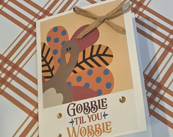 Puede incluir: Una tarjeta de felicitación con un pavo de dibujos animados con plumas estampadas en naranja, azul y marrón. La tarjeta incluye el texto GOBBLE TIL YOU WOBBLE, decorada con un lazo de rafia beige y pequeños adornos circulares dorados.