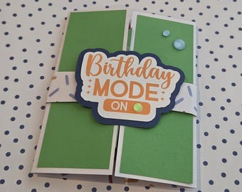 Puede incluir: Una tarjeta verde plegada con una banda blanca con patrón de chispas. Una etiqueta azul oscuro troquelada está centrada en la banda con el texto naranja Birthday MODE ON y pequeñas estrellas decorativas, acentuada por tres pequeñas gemas azul claro y verdes.