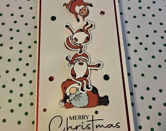 Puede incluir: Una tarjeta de felicitación rectangular con fondo blanco y borde de purpurina roja. La tarjeta muestra tres Papás Noel de dibujos animados apilados sobre un gnomo. El texto "MERRY Christmas" está impreso en negro en la parte inferior con gemas rojas y verde oscuro.