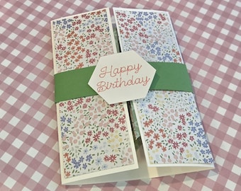 Puede incluir: Una tarjeta de felicitación plegada con un estampado floral de flores rojas, azules y rosas sobre fondo blanco. Una banda de papel verde liso envuelve el centro, sujeta por una etiqueta hexagonal blanca con el texto Happy Birthday en rojo.