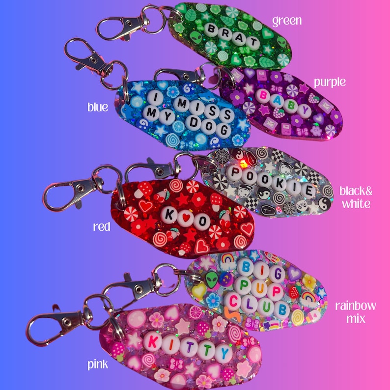 May include: Seven colorful keychains with different designs and text. The keychains are shaped like motel keys and are decorated with glitter, beads, and other embellishments. The text on the keychains includes "Brat", "I Miss My Dog", "Baby", "Pookie", "KO", "Big Pup Club", and "Kitty".