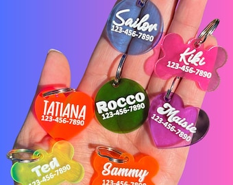 May include: A selection of brightly coloured, personalised pet ID tags in different shapes, including a heart, bone and circle. Each tag displays a name and phone number, such as "Sailor 123-456-7890", "Kiki 123-456-7890", and "Rocco 123-456-7890".