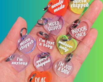 May include: A collection of 10 heart-shaped dog tags with various phrases and colors. The tags are made of clear acrylic and have white lettering. Some of the phrases include "micro chipped", "needs meds", "indoor only", "I'm just a baby", and "deaf".