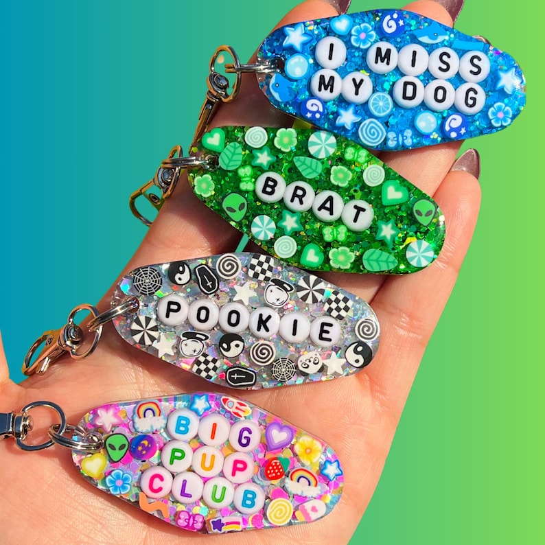 May include: Four colorful keychains with different words and designs. The keychains are oval shaped and have a silver metal ring. The words on the keychains are "I Miss My Dog", "Brat", "Pookie", and "Big Pup Club".