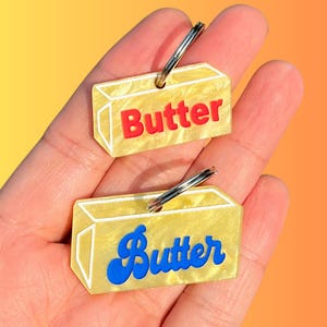 May include: Two butter-shaped keychains with silver key rings. The keychains are yellow with white outlines and the word "Butter" in red and blue. The keychains are held in a hand against a yellow and orange background.