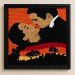 May include: A square framed crochet wall art piece depicting a couple kissing in silhouette against a vibrant orange sunset. The figures are rendered in black and beige yarn, with a red accent at the lips, set within a simple black frame.