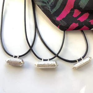 May include: Three silver pendant necklaces with black cords. Each pendant has a cylindrical shape with intricate designs. A colourful scarf with pink, black, and green patterns is in the background. The necklaces are displayed on a white surface.